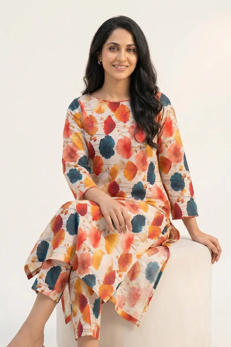 Model sitting on a white pouf wearing the Dorikhas Saffron Breeze 2 piece allover printed lawn dress in a vibrant saffron and teal floral print.