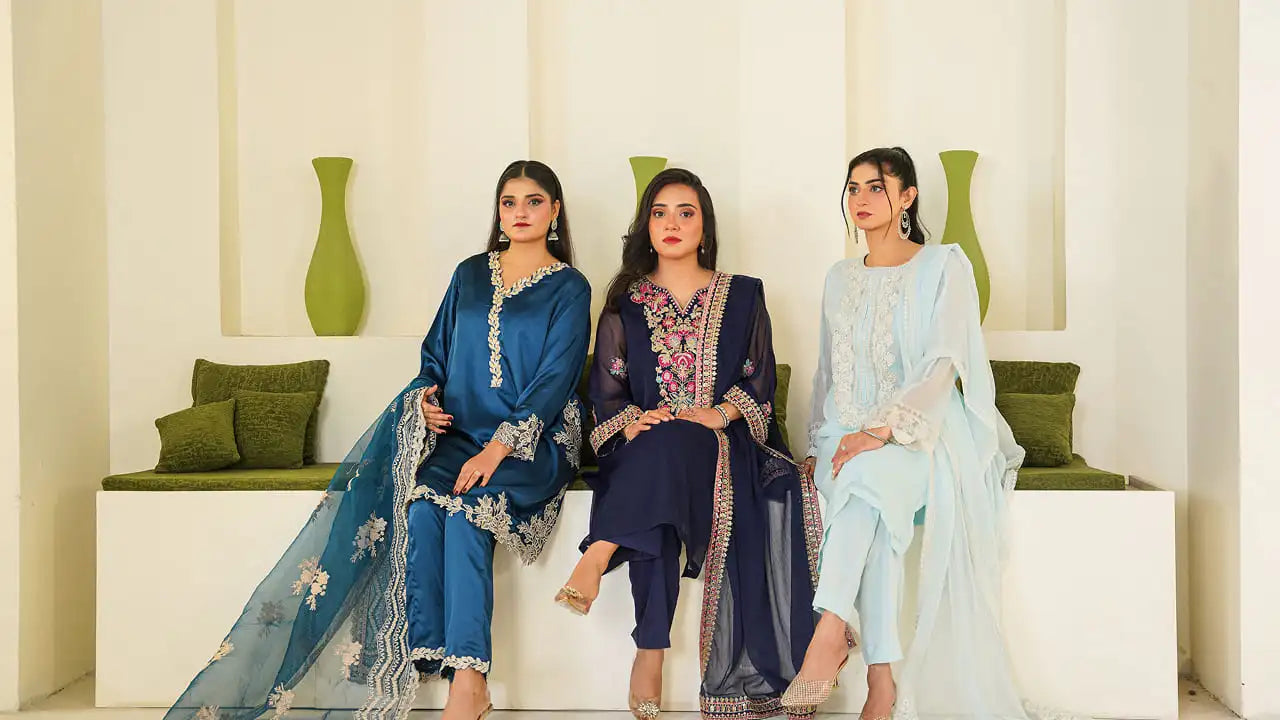 Three women sitting together showcasing Dorikhas premium women's apparel, featuring elegantly embroidered suits in deep teal, rich navy, and soft ice blue.
